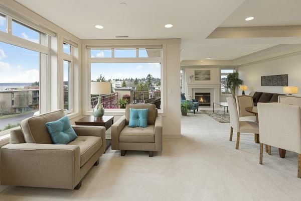 Bright, sunny living room with large windows, armchairs with teal pillows, and a light carpet