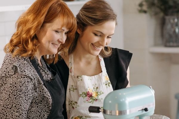Two women smiling over a mint-green stand mixer in a bright kitchen
