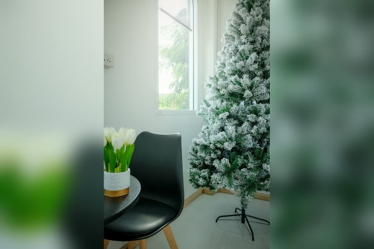 Snow-flocked Christmas tree on a metal stand beside a window, a black chair and white tulips (representative photo, not the exact product)
