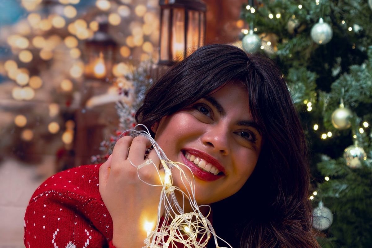 Smiling woman in a red holiday sweater holding a bundle of warm white Christmas lights by a tree