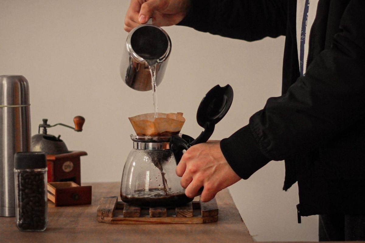 Hands pouring hot water over coffee grounds into a glass carafe next to a hand grinder and coffee beans