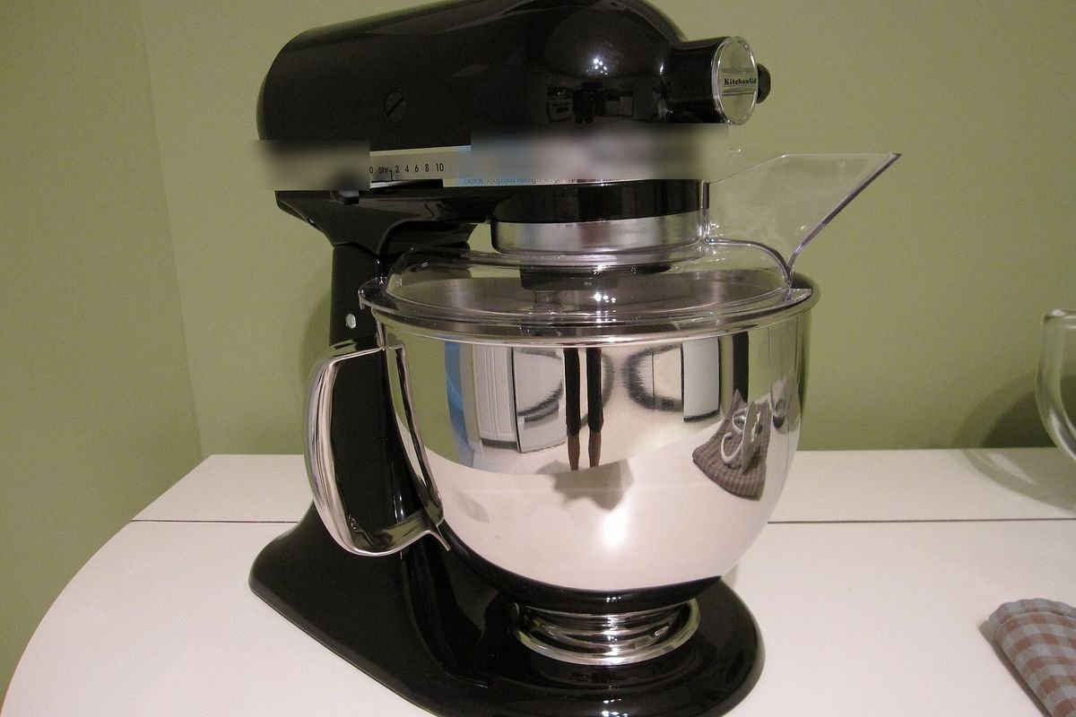 Black tilt-head stand mixer with a stainless bowl and clear pouring shield on a white counter (representative photo, not the exact product)