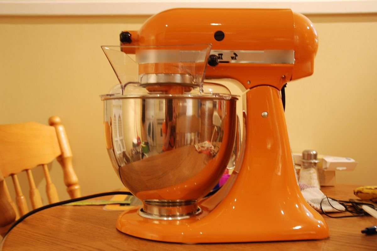 Orange tilt-head stand mixer with a stainless bowl on a wooden table (representative photo, not the exact product)