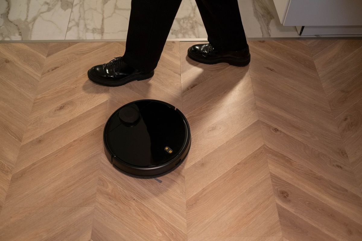 Black robot vacuum on a herringbone floor as a person walks past (representative photo, not the exact product)