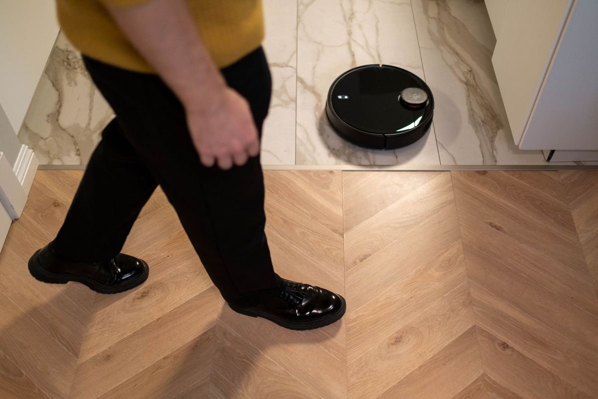 Black robot vacuum cleaning near a person walking across a wood floor (representative photo, not the exact product)