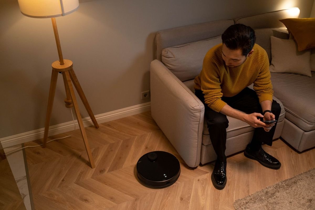 Man on a sofa using his phone while a robot cleaner runs on the living room floor (representative photo, not the exact product)